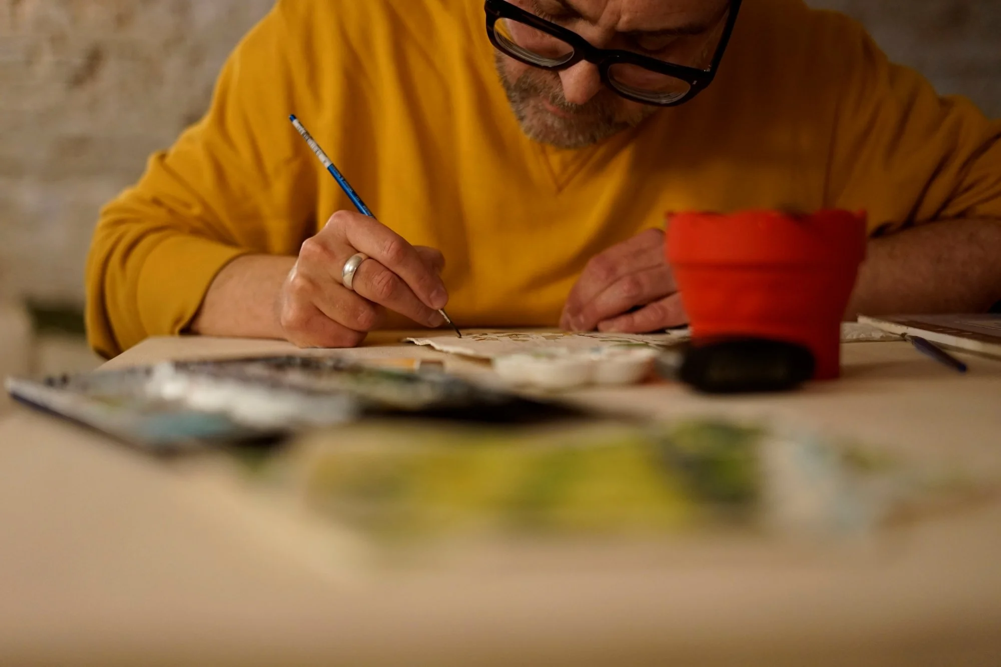 A man with glasses and a gray beard wearing a yellow shirt, sitting at a table and painting or drawing with a small brush on paper. There is a red container and art supplies on the table.