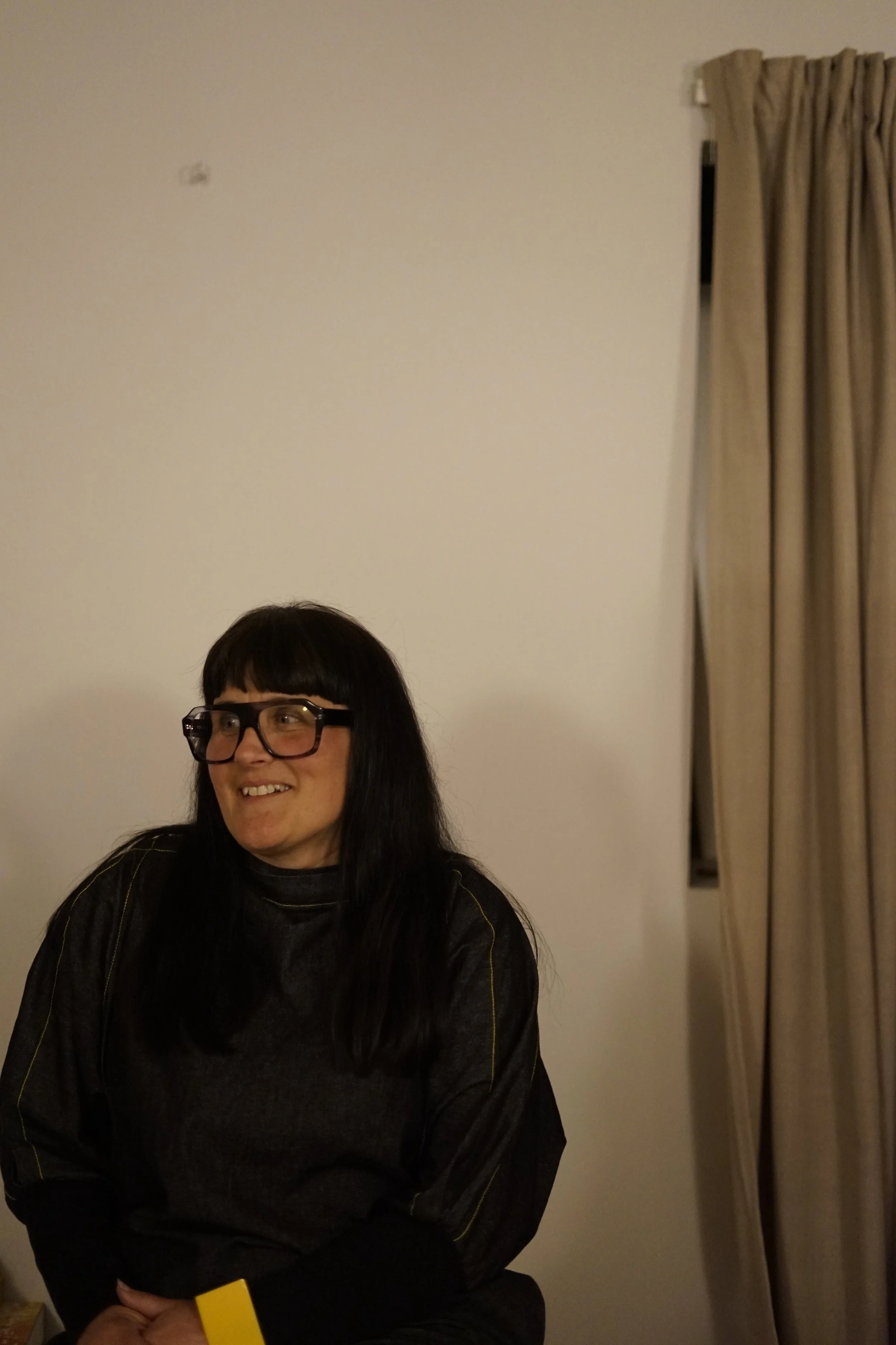 A person with long black hair, glasses, and a dark top, smiling and sitting indoors near a plain wall and beige curtains.