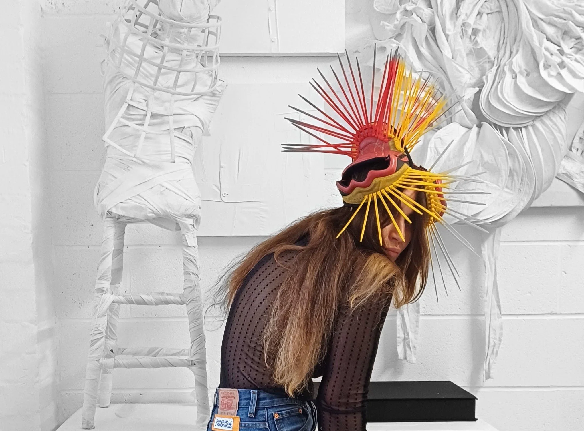 A woman wearing an avant-garde mask with multicolored spikes in an art studio with white sculptures and artwork in the background.