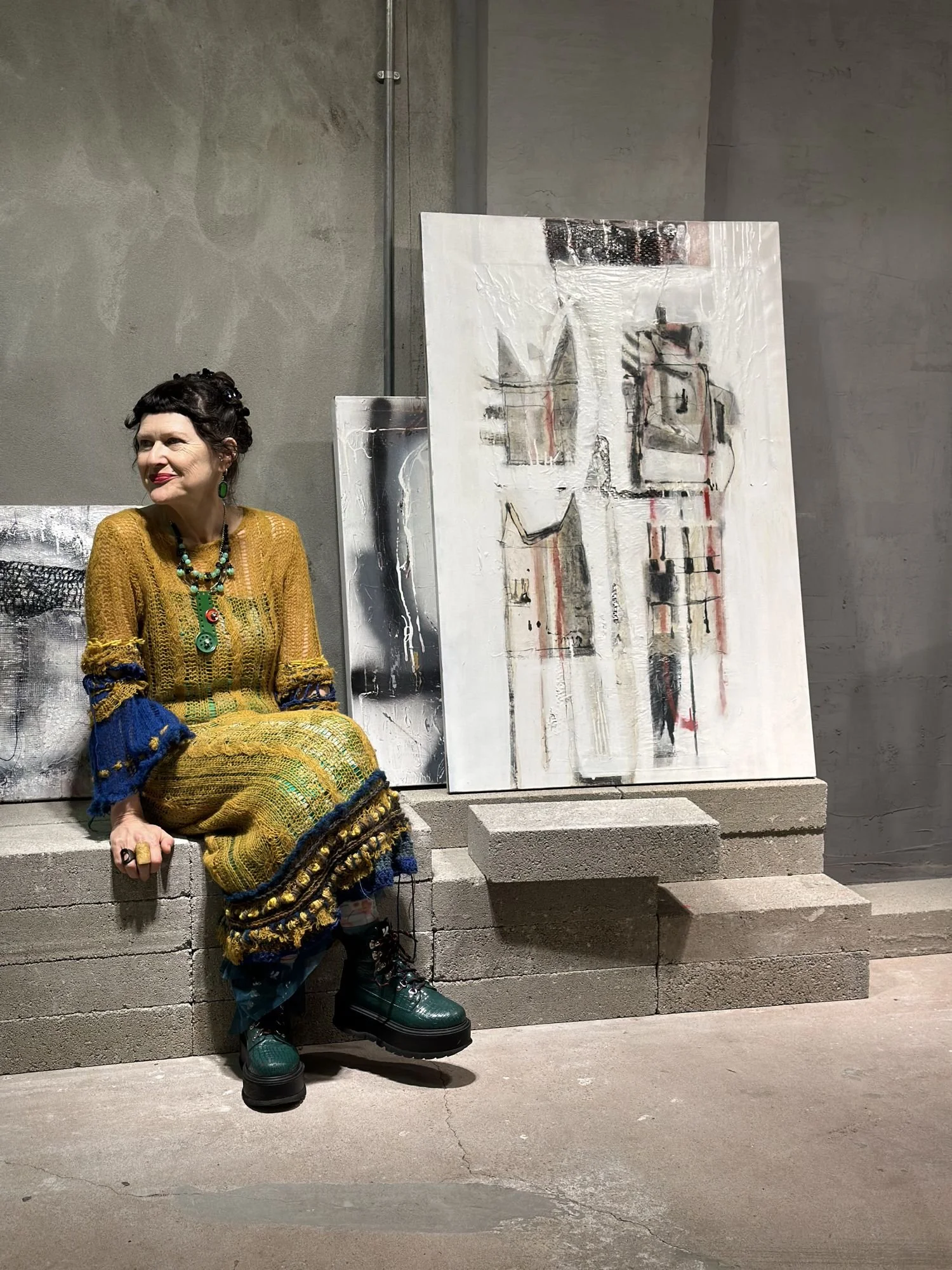 Woman dressed in colorful knit dress sitting on concrete steps beside standing abstract paintings in an art gallery.
