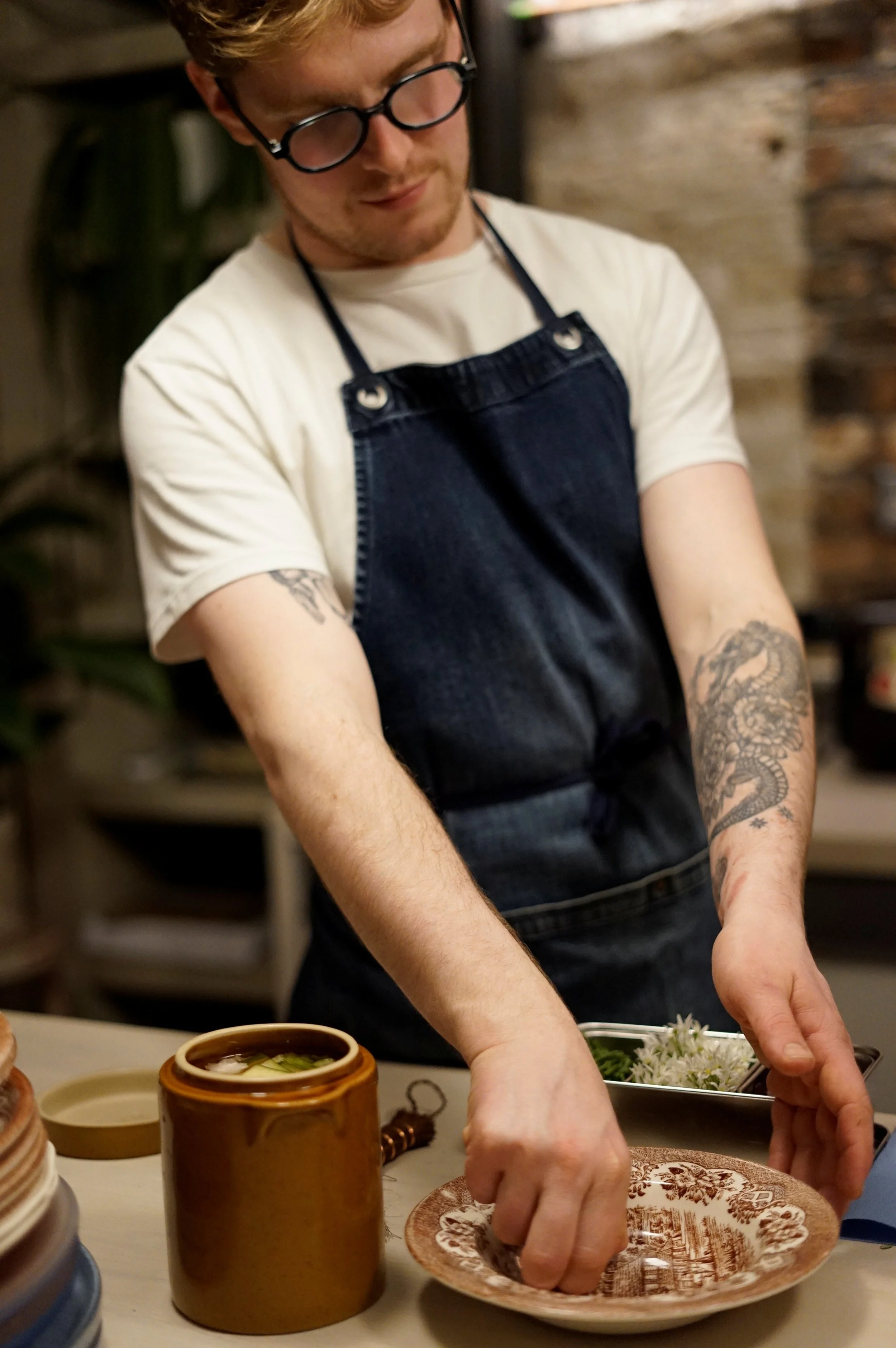 A man with glasses and tattoos wearing a white t-shirt and a black apron arranging dishes on a table in a rustic kitchen.