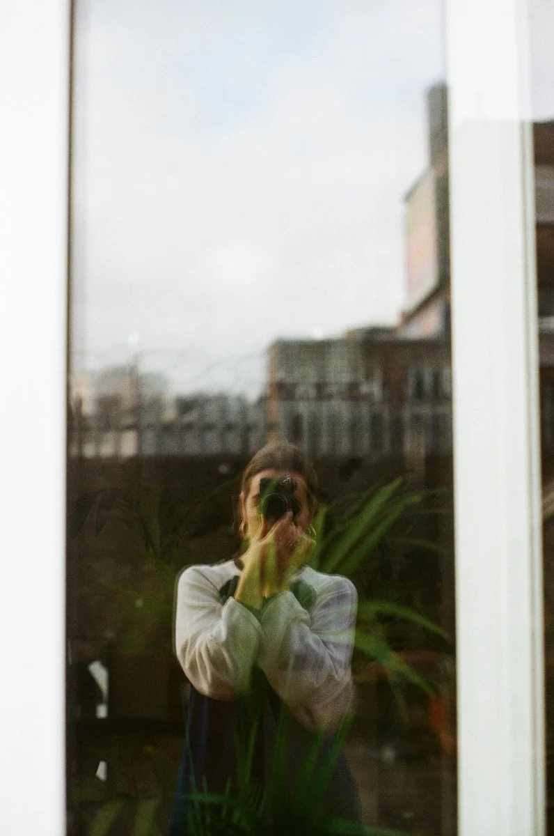 Person taking a photo of their reflection in a glass door or window, with buildings and plants visible outside.