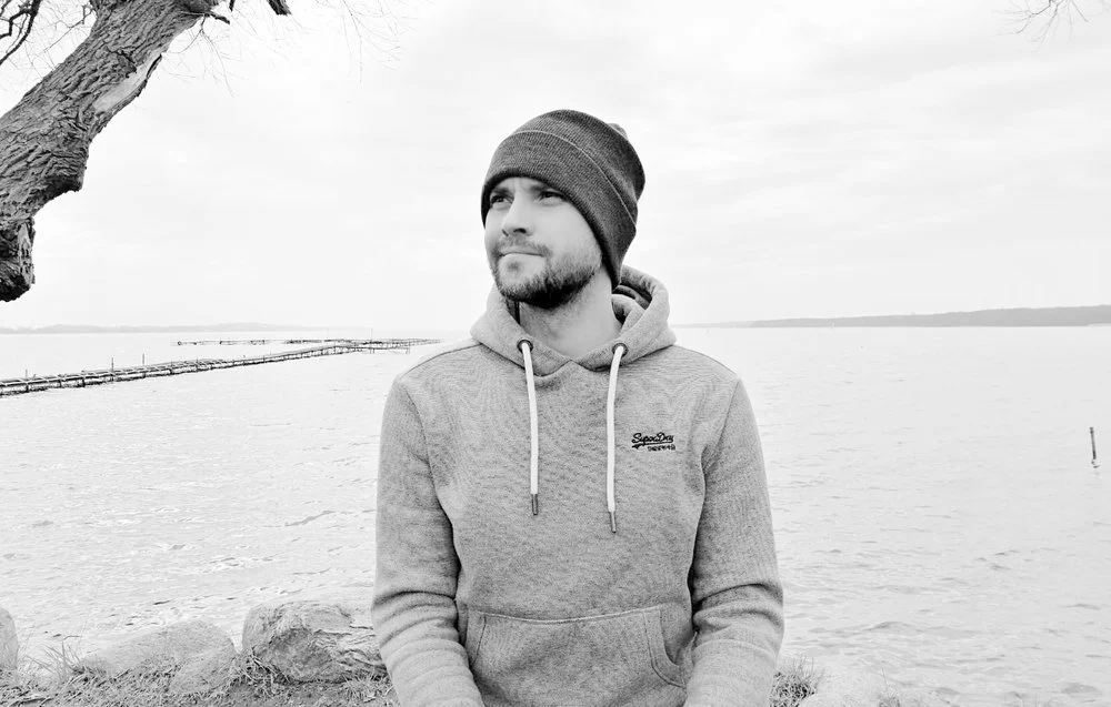 A man in a hoodie and beanie sitting outdoors near a body of water, with a tree branch overhead and a dock in the background.