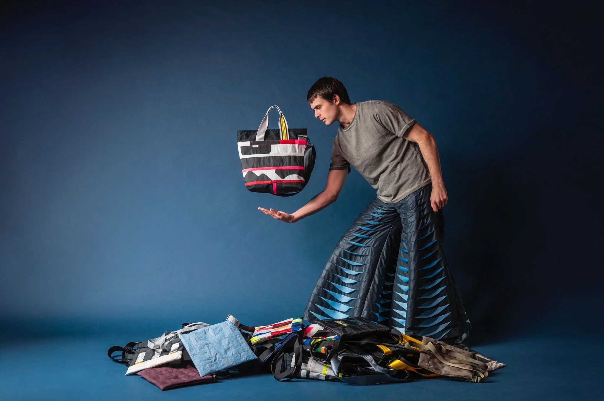 A man in a gray T-shirt and patterned pants appears to be balancing a prepared shopping or travel bag in mid-air, with various travel bags, towels, and accessories scattered on the floor around him against a dark blue background.