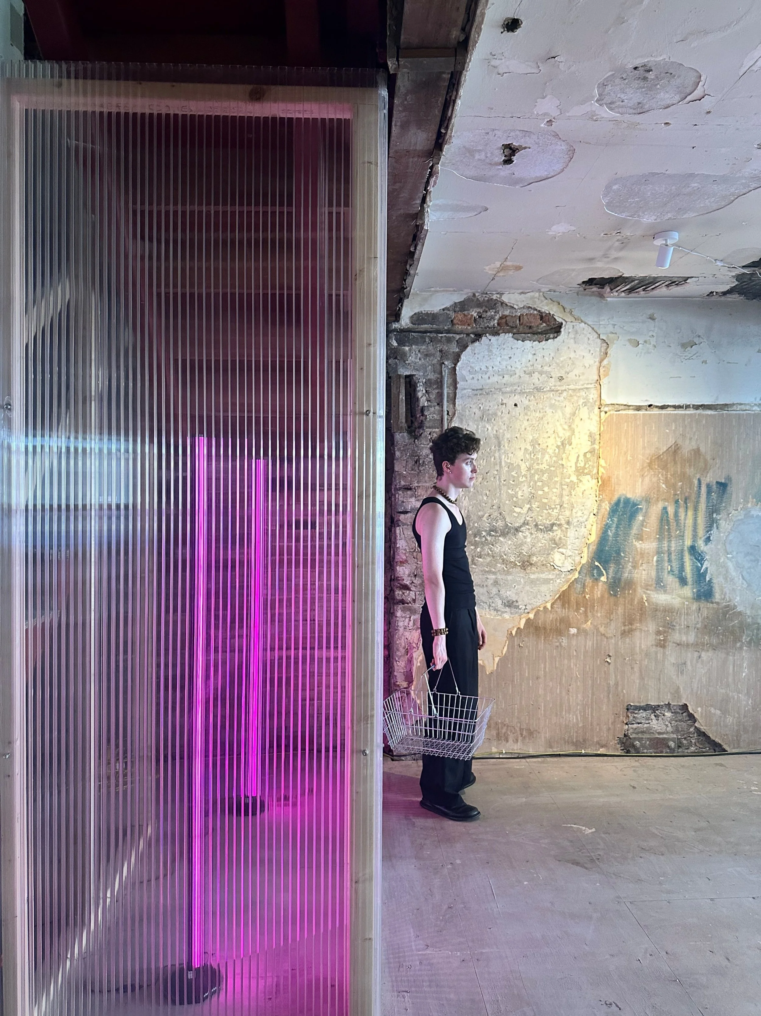 A woman with a short hairstyle dressed in black, holding a wire shopping basket, standing in front of a grunge wall with peeling paint and exposed brick in an industrial setting. There is a neon pink light on the left side.