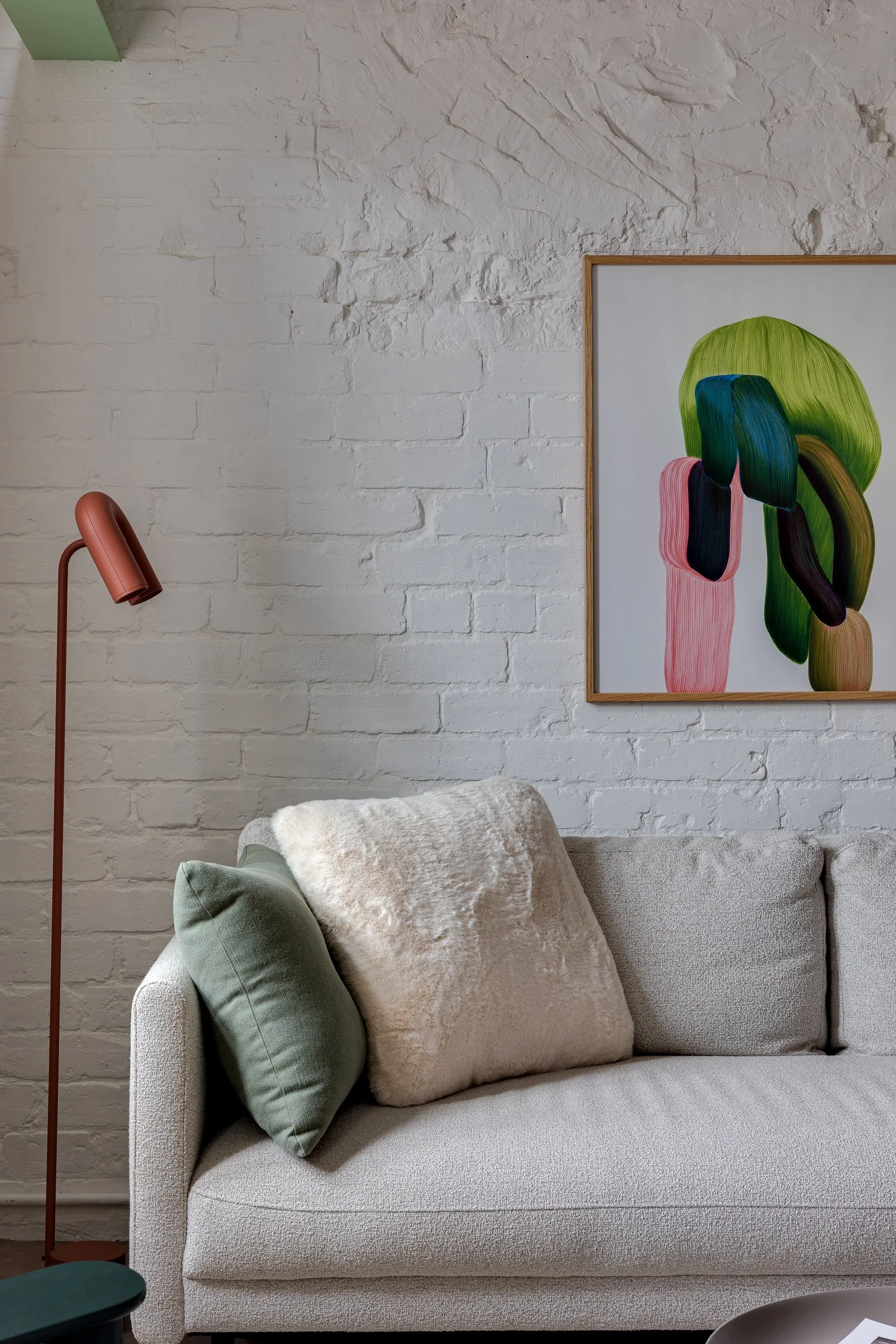 A modern living room corner with a white sofa, green and beige pillows, a tall rust-colored floor lamp, a white brick wall, and an abstract colorful artwork hanging on the wall.