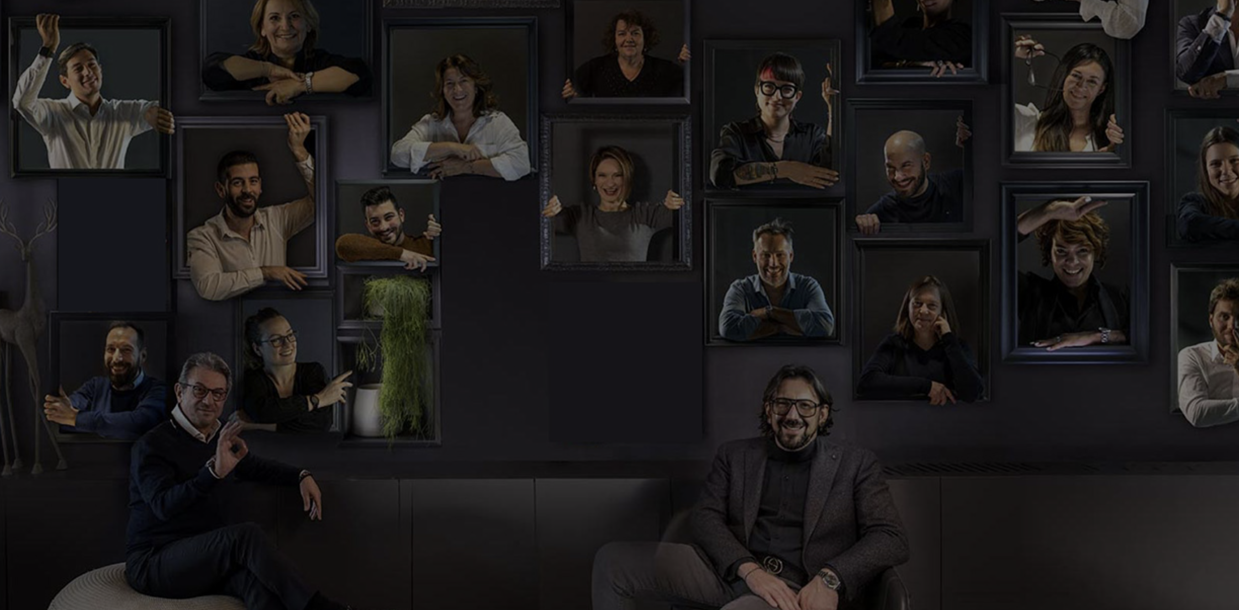 Group of people, including men and women, positioned within and around picture frames on a dark wall, with some frames empty. The individuals are smiling and pretending to interact with those in the frames, creating a playful and creative group photo.