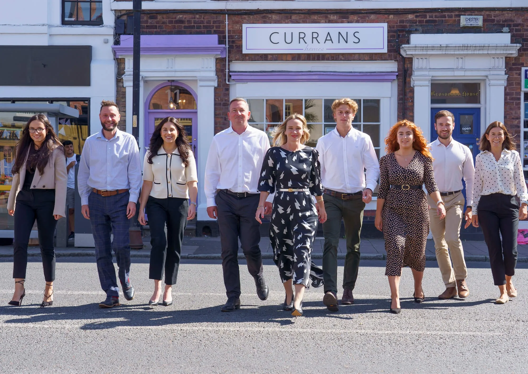 A diverse group of nine professionals walking confidently across the street in business casual attire, smiling, with a row of storefronts behind them, including a store named 'Currans Homes'.