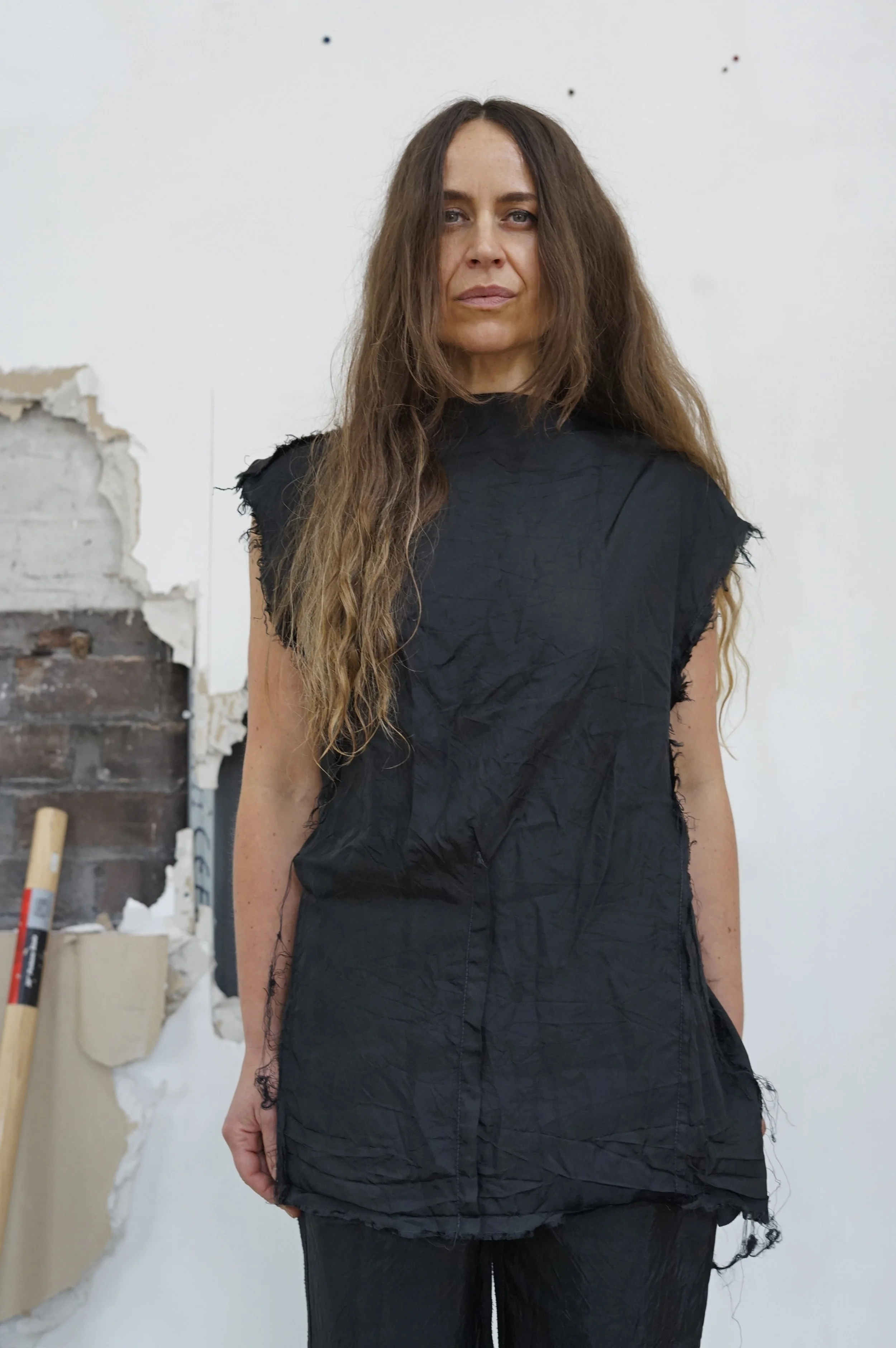 A woman with long, wavy brown hair standing in front of a partially demolished wall, wearing a black sleeveless top and black pants.