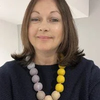 Close-up of a woman with shoulder-length brown hair wearing a black top, a navy blue sweater, and a colorful beaded necklace, smiling lightly.