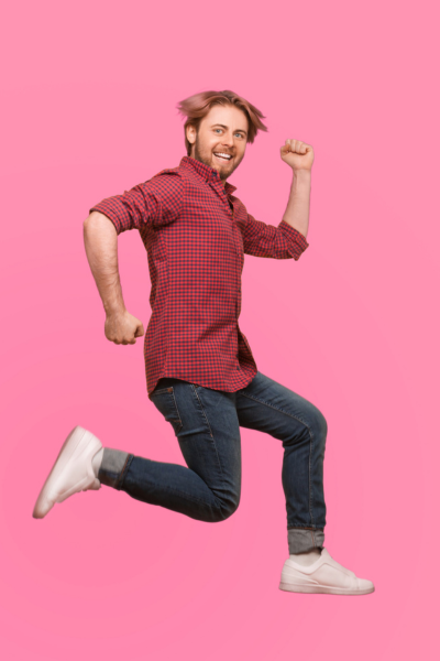 Un homme souriant, vêtu d'une chemise à carreaux rouges, sautant devant un fond rose