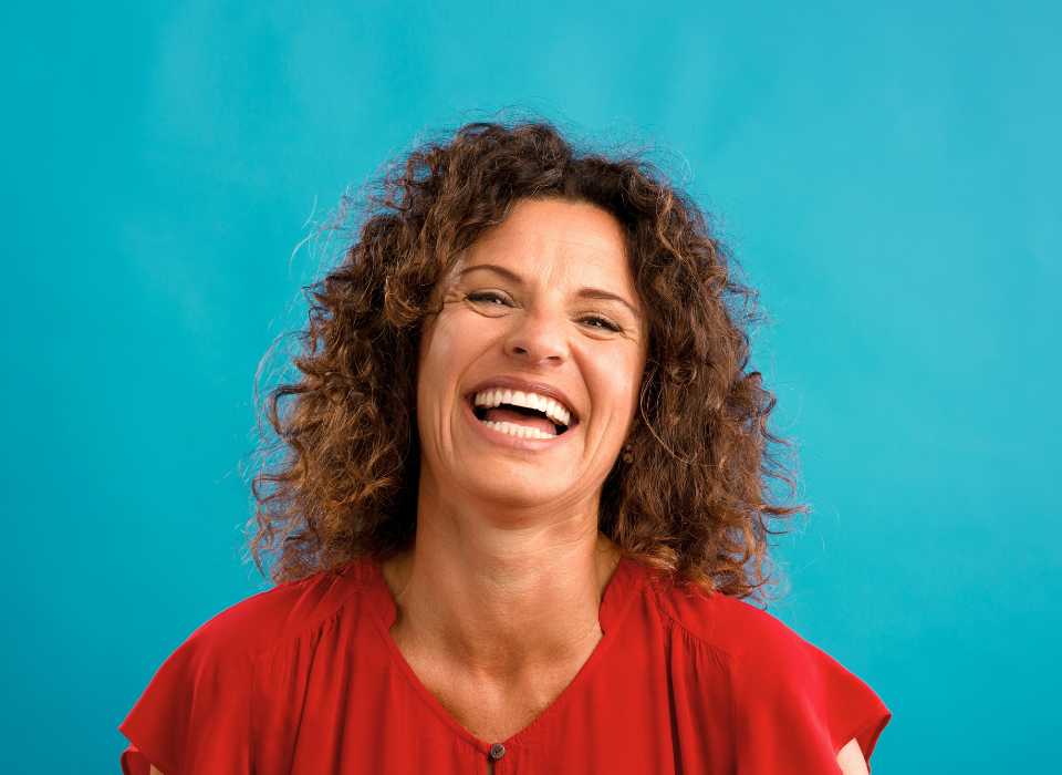 Femme souriante avec des cheveux bouclés, portant une chemise rouge, devant un fond bleu.