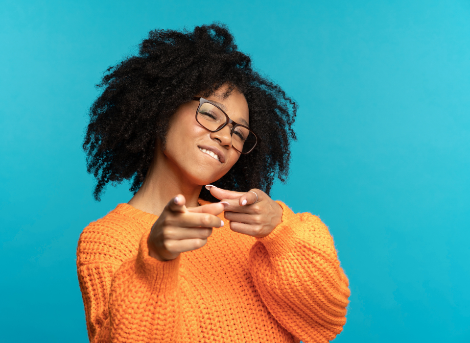 Jeune femme noire avec des cheveux bouclés, portant des lunettes et un pull orange, souriant et pointant ses doigts vers l'objectif, devant un fond bleu.