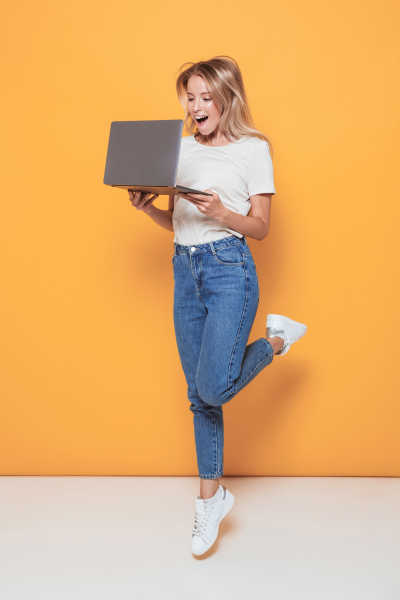 Jeune femme souriante debout sur un pied, tenant un ordinateur portable, contre un fond jaune