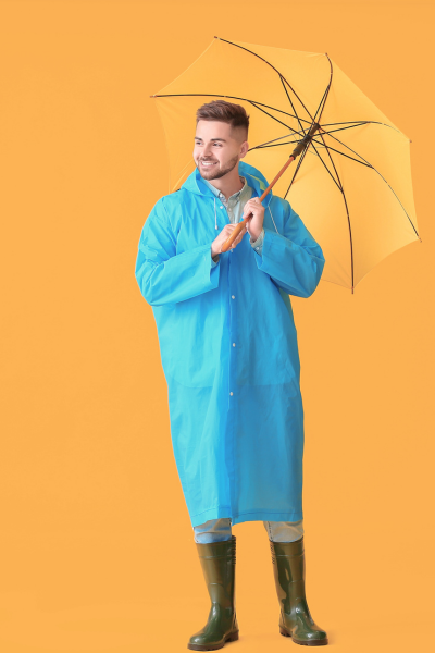 Homme souriant portant un imperméable bleu, des bottes de caoutchouc noires et tenant un parapluie jaune contre un fond orange.