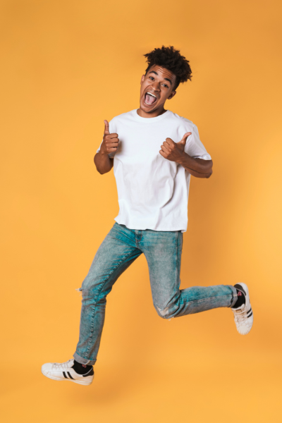 Jeune homme souriant, sautant avec le pouce en l'air, portant un t-shirt blanc et un jean, sur un fond orange.