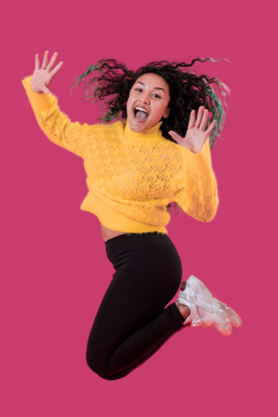 Jeune femme souriante sautant en l'air, portant un pull jaune, un pantalon noir, et des baskets blanches, avec un fond rose.