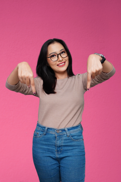 Femme souriante portant des lunettes, un pull beige et un jean bleu, pointant vers le bas avec les pouces, devant un fond rose.