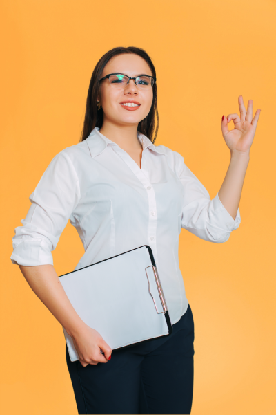 Femme souriante portant des lunettes, une chemise blanche et tenant un classeur, faisant le geste de l'ok avec la main gauche devant un fond jaune.