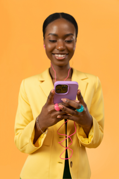 Femme souriante regardant son téléphone avec des écouteurs, portant un blazer jaune, fond orange.