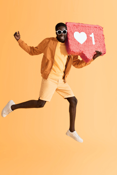 Un homme souriant en lunettes de soleil, portant une veste orange, un t-shirt beige, un short beige, et des baskets blanches, sautant avec un grand symbole de publication de réseaux sociaux en forme de cœur et d'une mention «1».