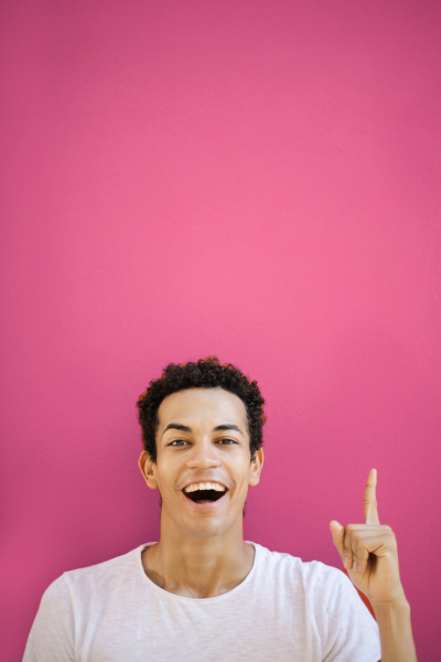 Jeune homme souriant portant un t-shirt blanc, pointant vers le haut avec le doigt, fond rose.