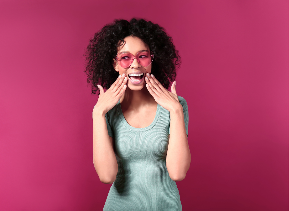 Jeune femme souriante portant des lunettes roses, avec des cheveux bouclés, sur un fond rose