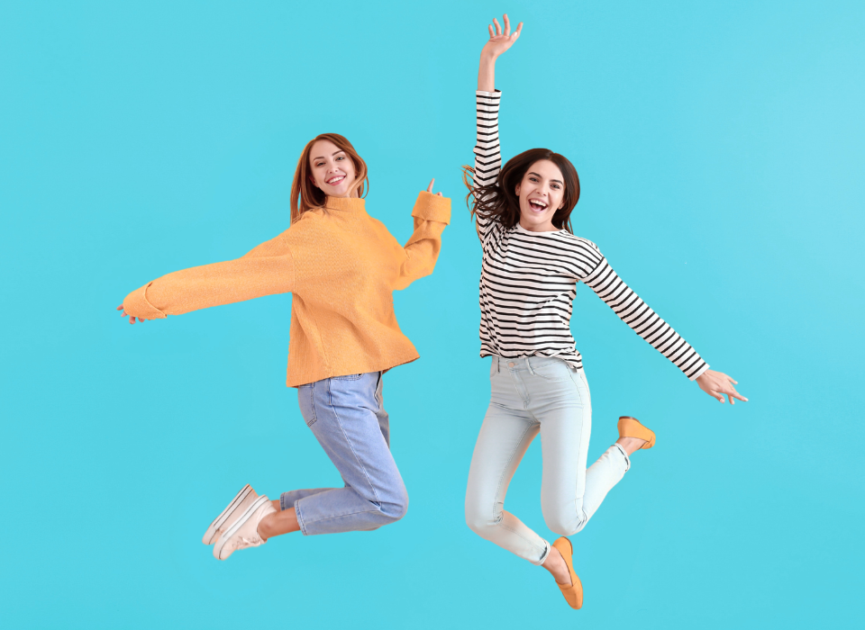 Deux jeunes femmes souriantes sautent en l'air sur un fond bleu, habillées de vêtements décontractés.