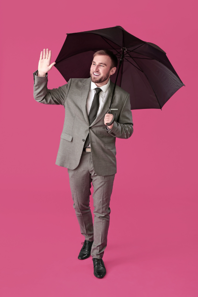 Homme en costume gris tenant un parapluie noir et faisant un signe de la main avec un sourire, sur fond rose.