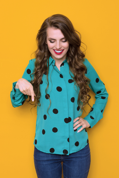 Femme souriante pointant vers le bas, portant une chemise turquoise à pois noirs, contre un fond jaune.