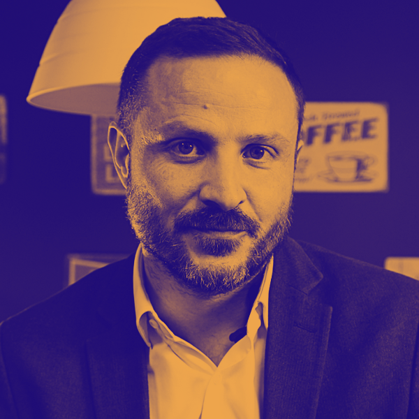 Un homme avec une barbe, portant un costume, dans un intérieur avec une enseigne 'Café' en arrière-plan.