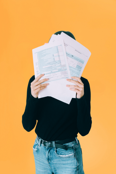 Personne tenant plusieurs papiers ou documents, se cachant le visage, contre un fond orange