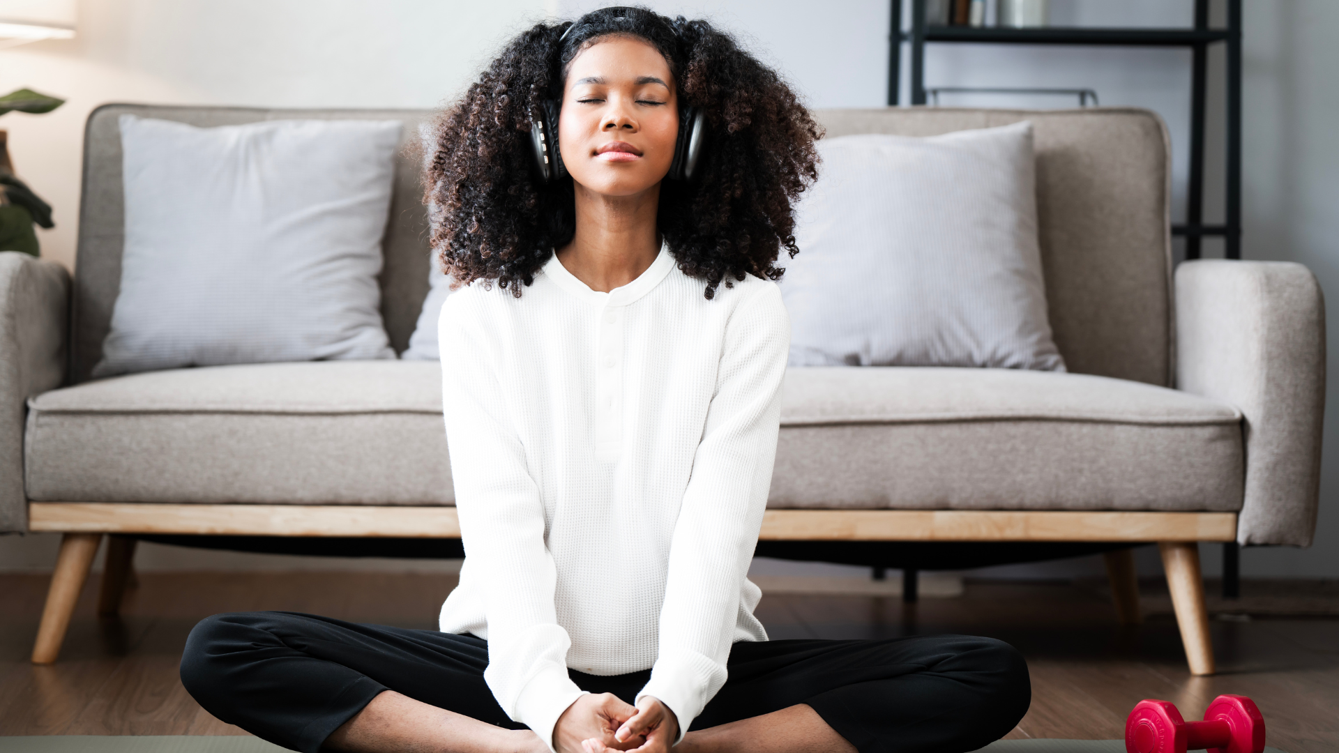 A woman with curly hair and headphones sitting cross-legged on the floor with eyes closed, in a living room with a sofa and a dumbbell nearby.