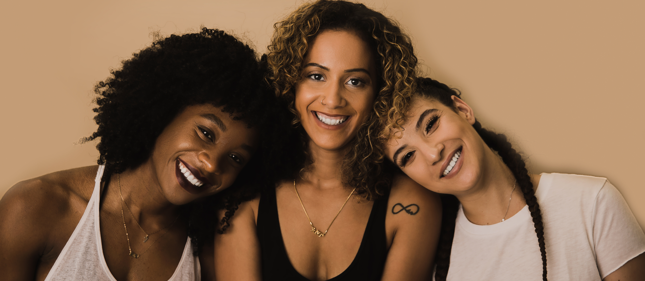 Three women smiling together, closely positioned against a beige background.