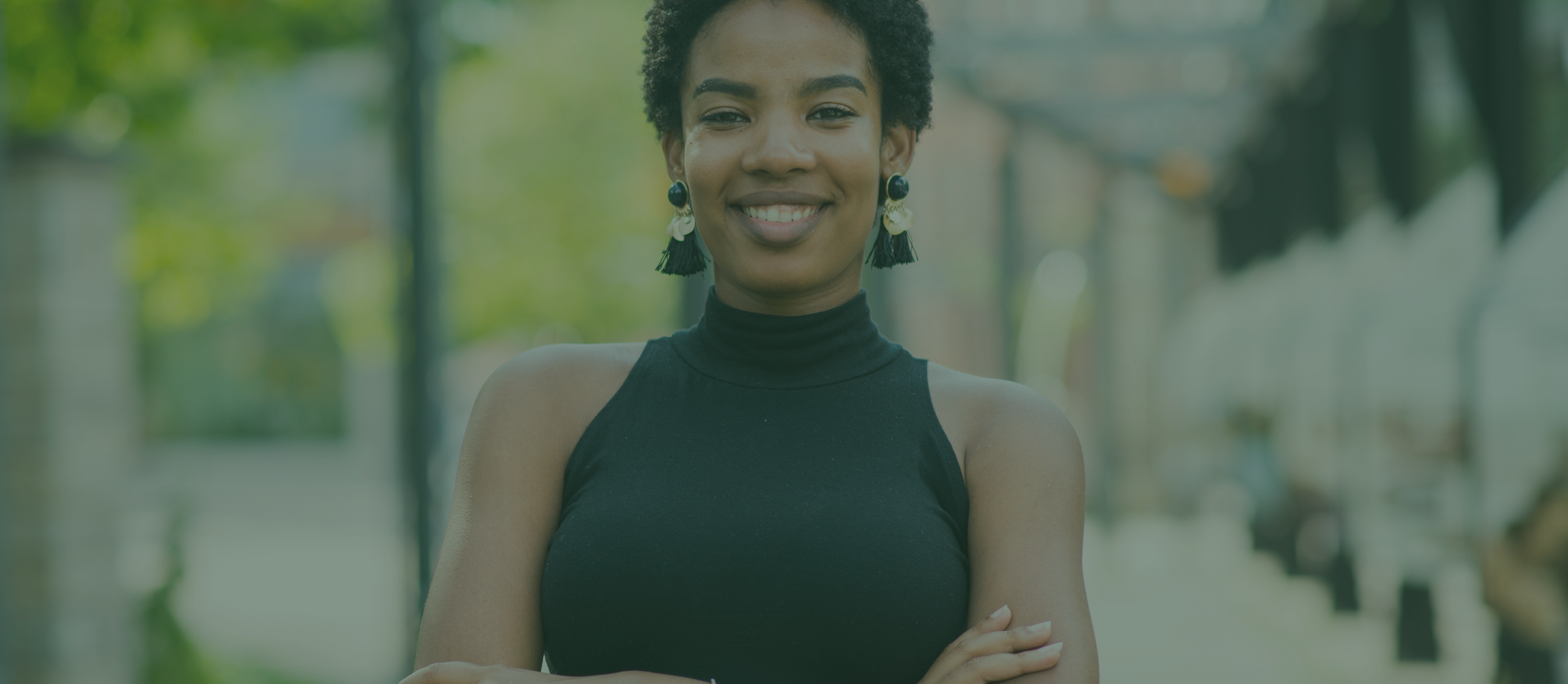 Portrait of a smiling young African American woman with short curly hair, wearing a sleeveless black turtleneck top and colorful earrings, standing outdoors in a green, blurry background.