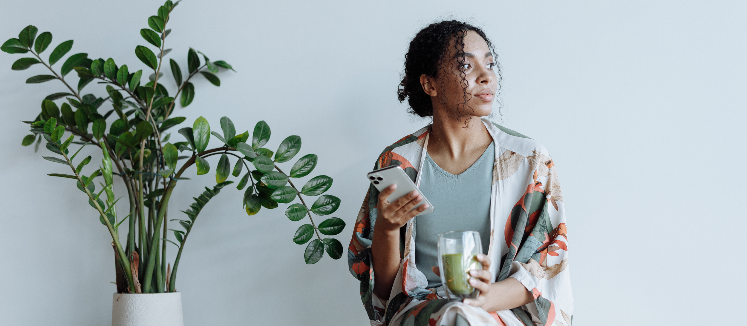 A young woman sitting indoors holding a smartphone in one hand and a glass of green juice in the other, looking to her right. Next to her is a large potted green plant against a light-colored wall.