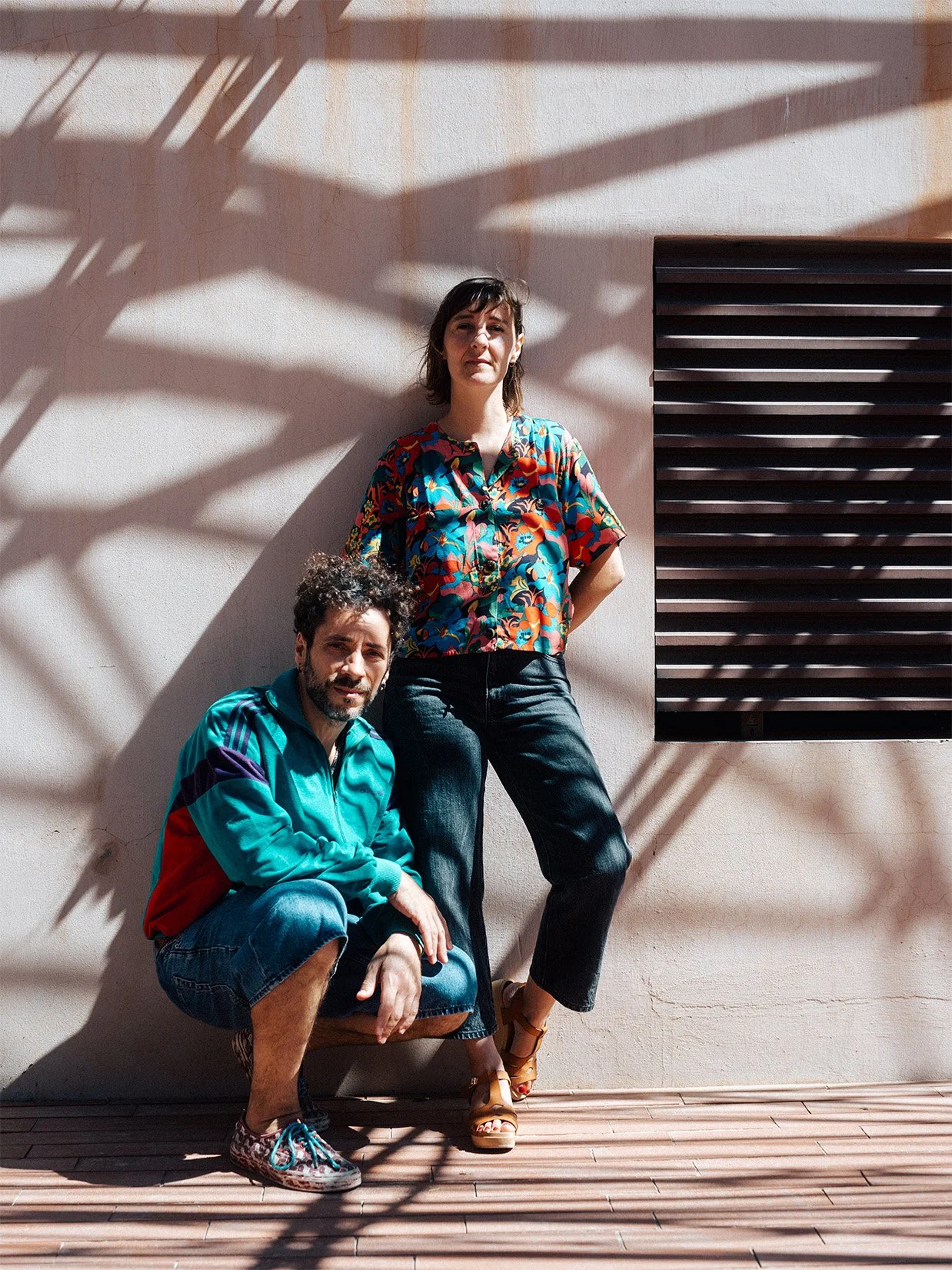 Deux personnes posent devant un mur au soleil, avec des ombres de plantes et de structures sur le mur et le sol. La femme se tient debout avec une expression neutre, portant une chemise colorée, tandis que l'homme est assis en crouch avec une expression souriante, portant une veste bleu avec des détails colorés.