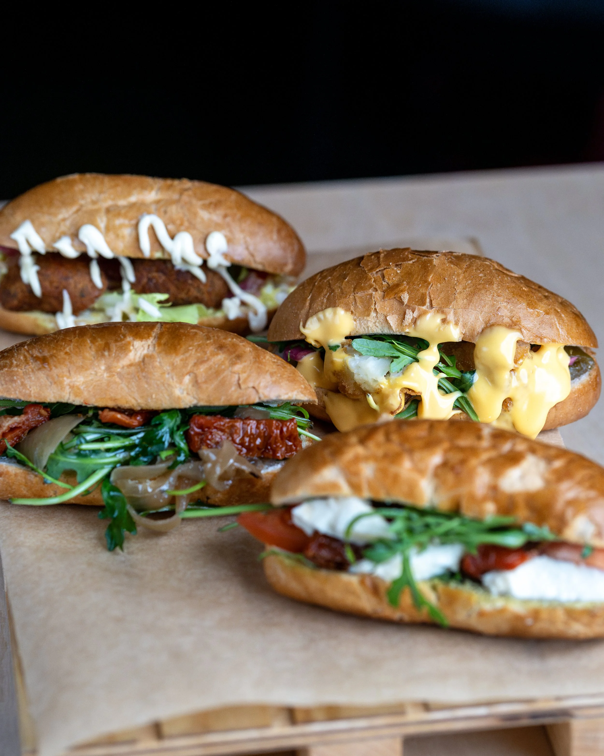 Four different gourmet sandwiches Montaditos, on a wooden tray with parchment paper. A fried chicken sandwich, a vegetarian sandwich, and a Caprese sandwich with mozzarella, tomato, and basil. Riga, Latvia, bar, dining, snacks, pub