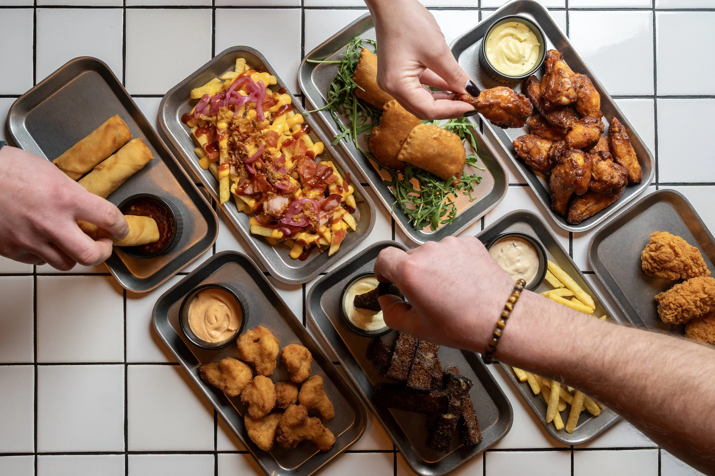Several small metal trays of various appetizers and fried foods on a white tiled table, with hands holding snacks and dipping sauces. Rīga, Riga, Gastropub, bar, restaurant, dining, nightlife in Latvia