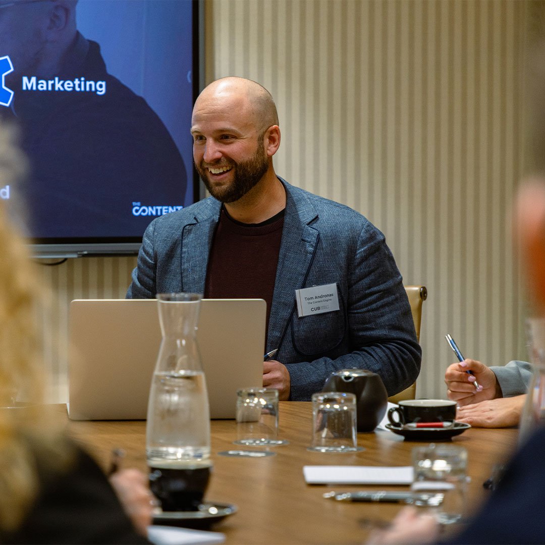 A man with a beard in a gray blazer and a dark shirt sitting at a conference table with a laptop, glass water pitcher, cups, and notebooks, smiling during a meeting. In the background, a screen displays the words 'Marketing' and 'the CONTENT'.
