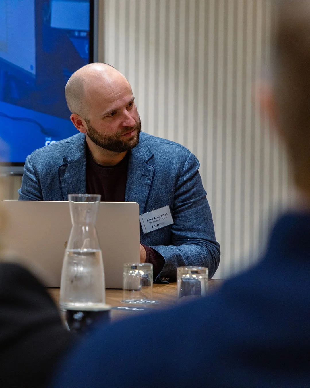 A man with a beard and a bald head, wearing a blue blazer, sitting at a table with a laptop, a water pitcher, and glasses, looking to his left during a meeting or conference.