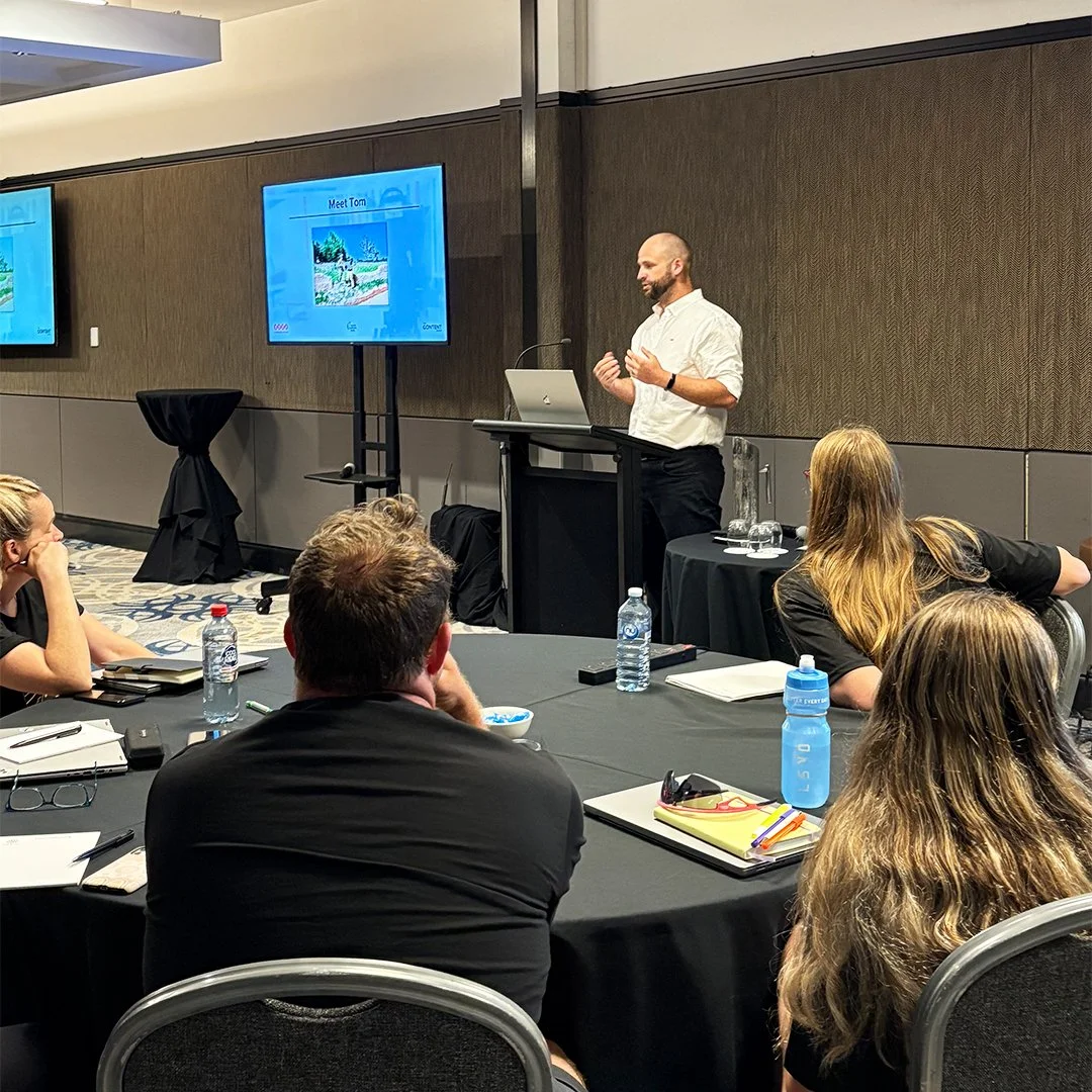 A man giving a presentation to a group of people sitting at a conference table in a hotel or conference room.
