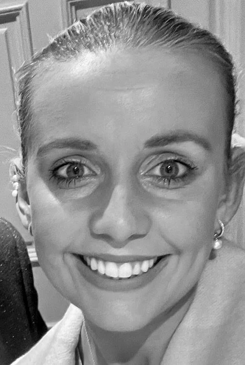 Close-up of a smiling woman with light-colored hair pulled back, wearing earrings, in a black and white photo.