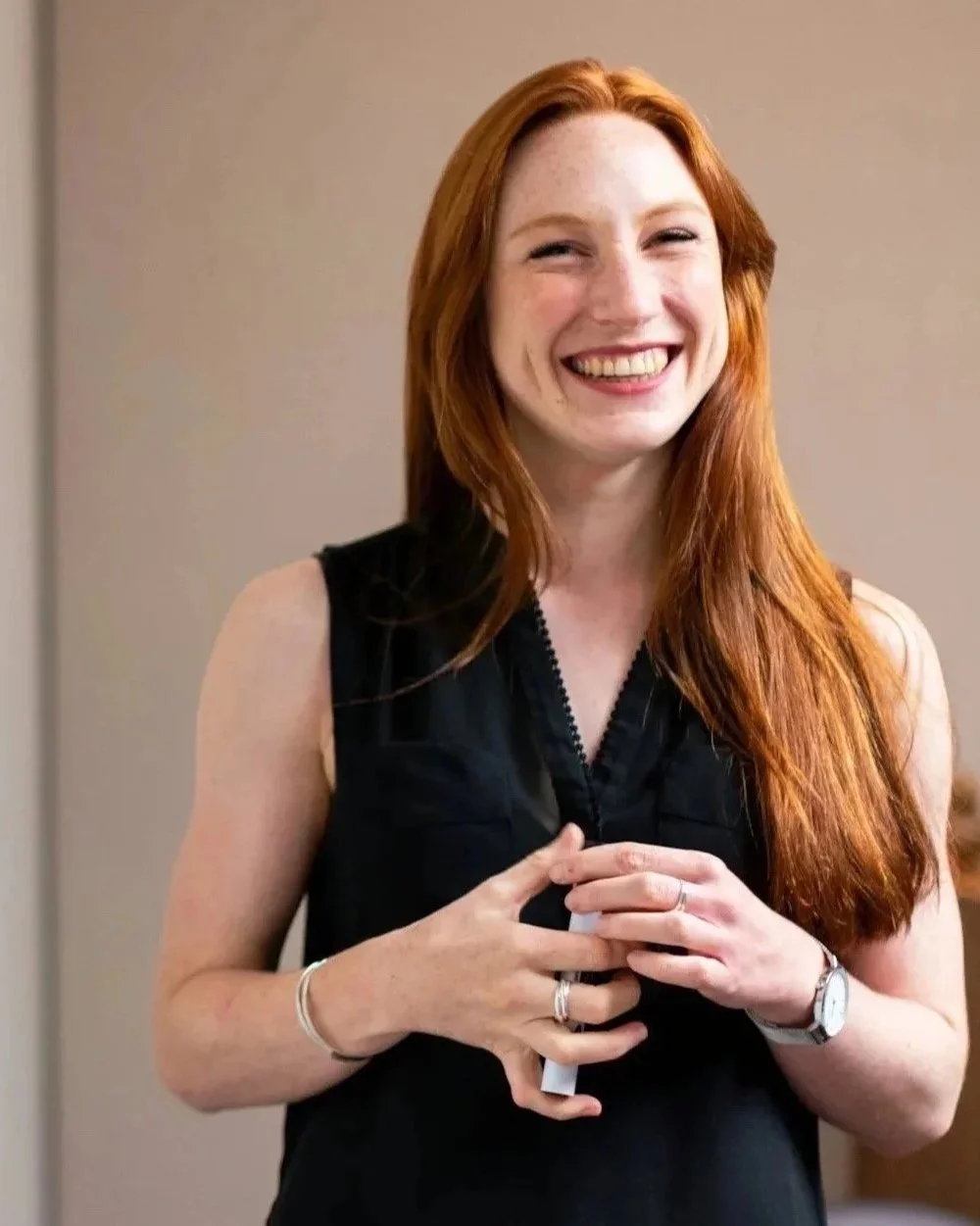 A smiling woman with long red hair wearing a black sleeveless top, holding a cup, standing against a neutral background.