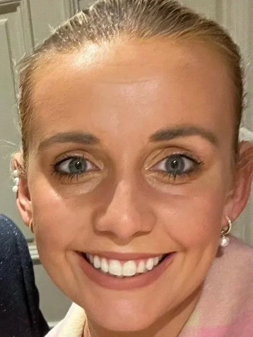 Close-up of a woman smiling, with blonde hair pulled back, blue eyes, and wearing pearl earrings.