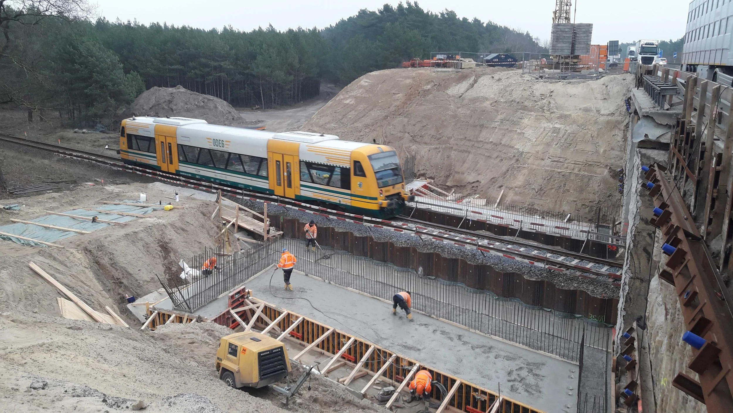 Betonagearbeiten am Bauwerk BW12 der A24 mit Bauarbeitern in der Baugrube und vorbeifahrendem ODEG-Zug auf der Bahnstrecke.
