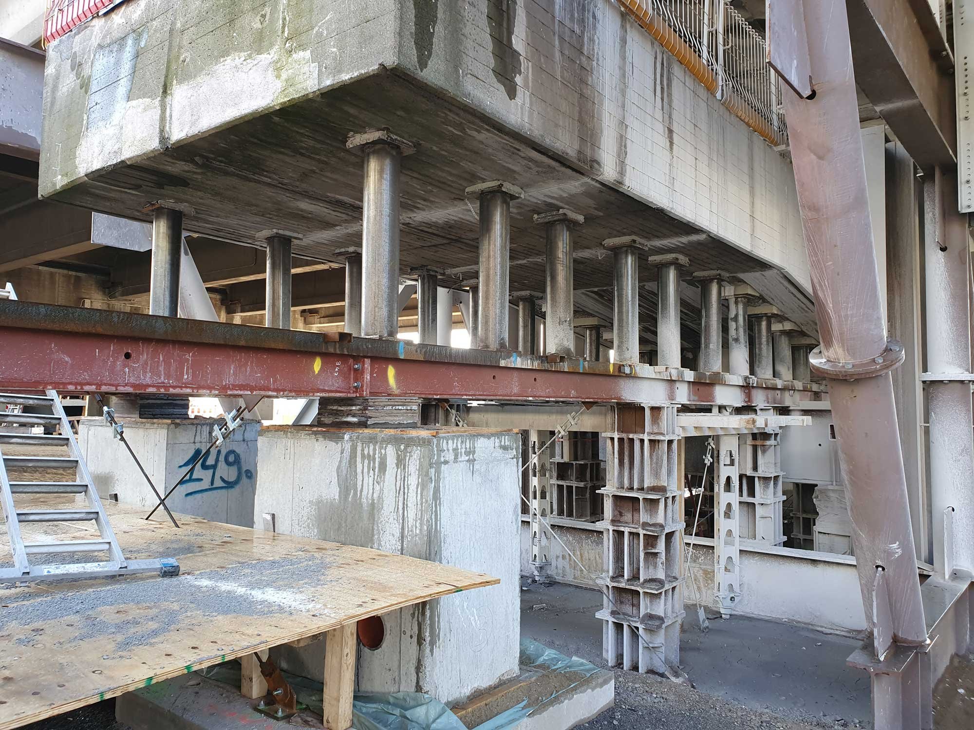 Unterseite einer Brücke während Bau- oder Sanierungsarbeiten: Ein massiver Betonträger wird durch zahlreiche Stahlstützen und Träger abgestützt, mit sichtbaren Schalungen und Bauelementen.