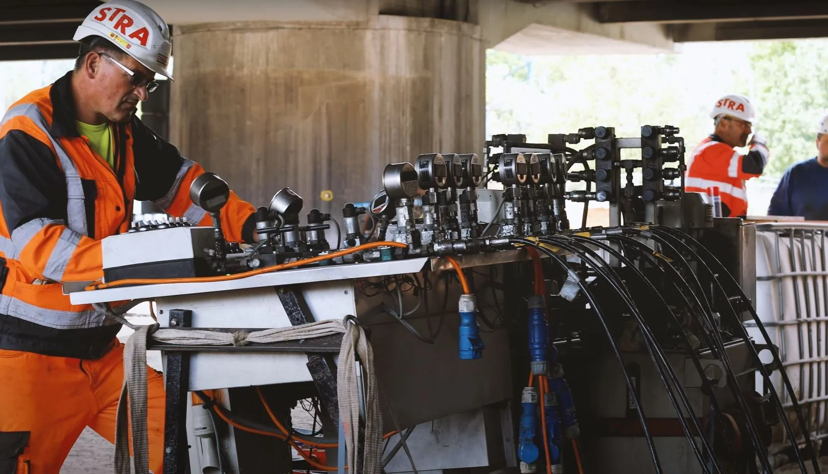 Ein Arbeiter in orangefarbener Schutzkleidung und mit weißem Schutzhelm bedient eine technische Anlage mit mehreren Ventilen, Anzeigen und Kabeln. Im Hintergrund stehen weitere Arbeiter unter einer Betonbrücke oder in einer Industrieumgebung.