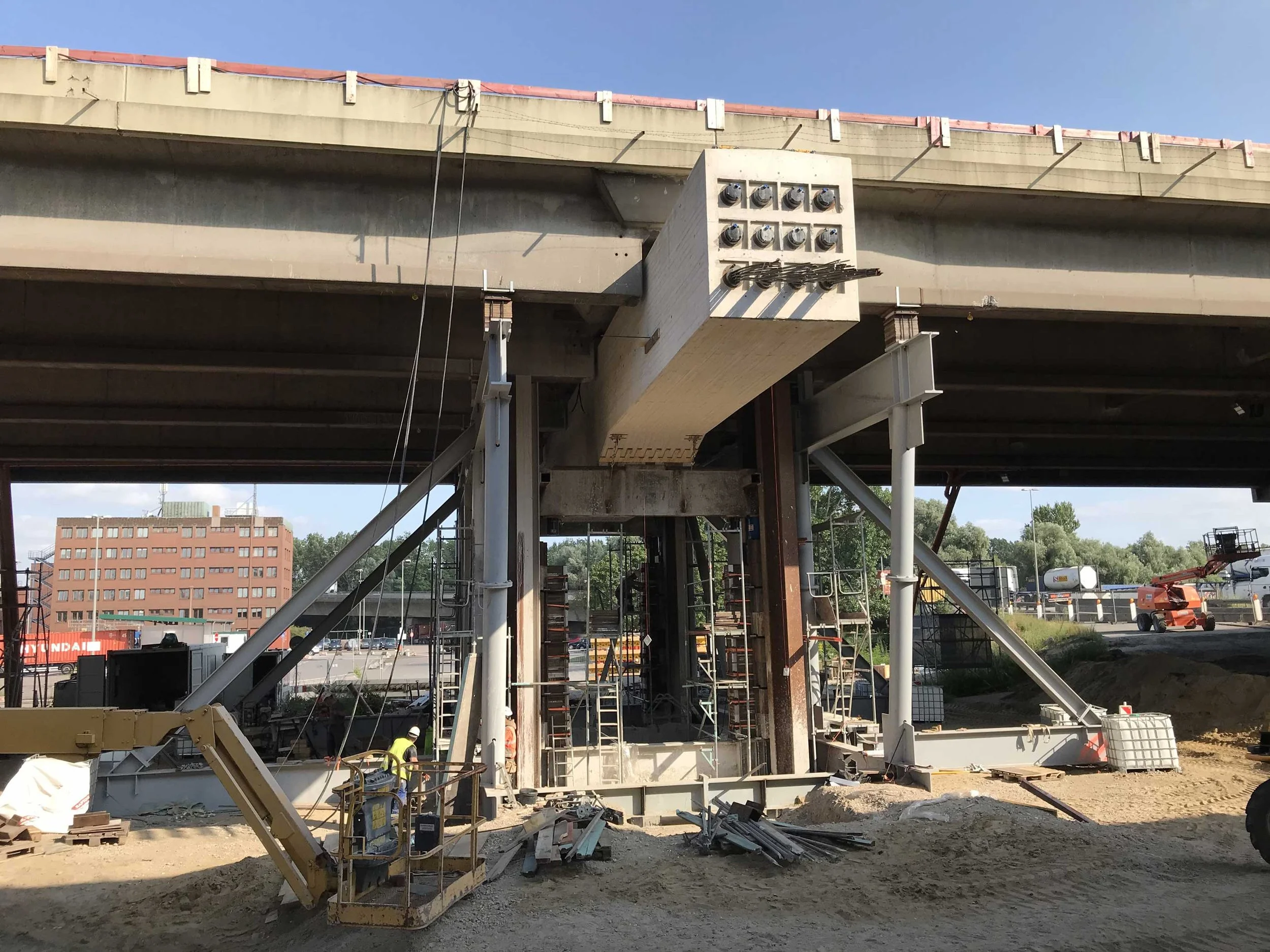 Baustelle unter einer Brücke mit Stahlstützen und Schalung: Ein großer Betonträger wird abgestützt und bearbeitet, während Arbeiter und Baugeräte im Einsatz sind.