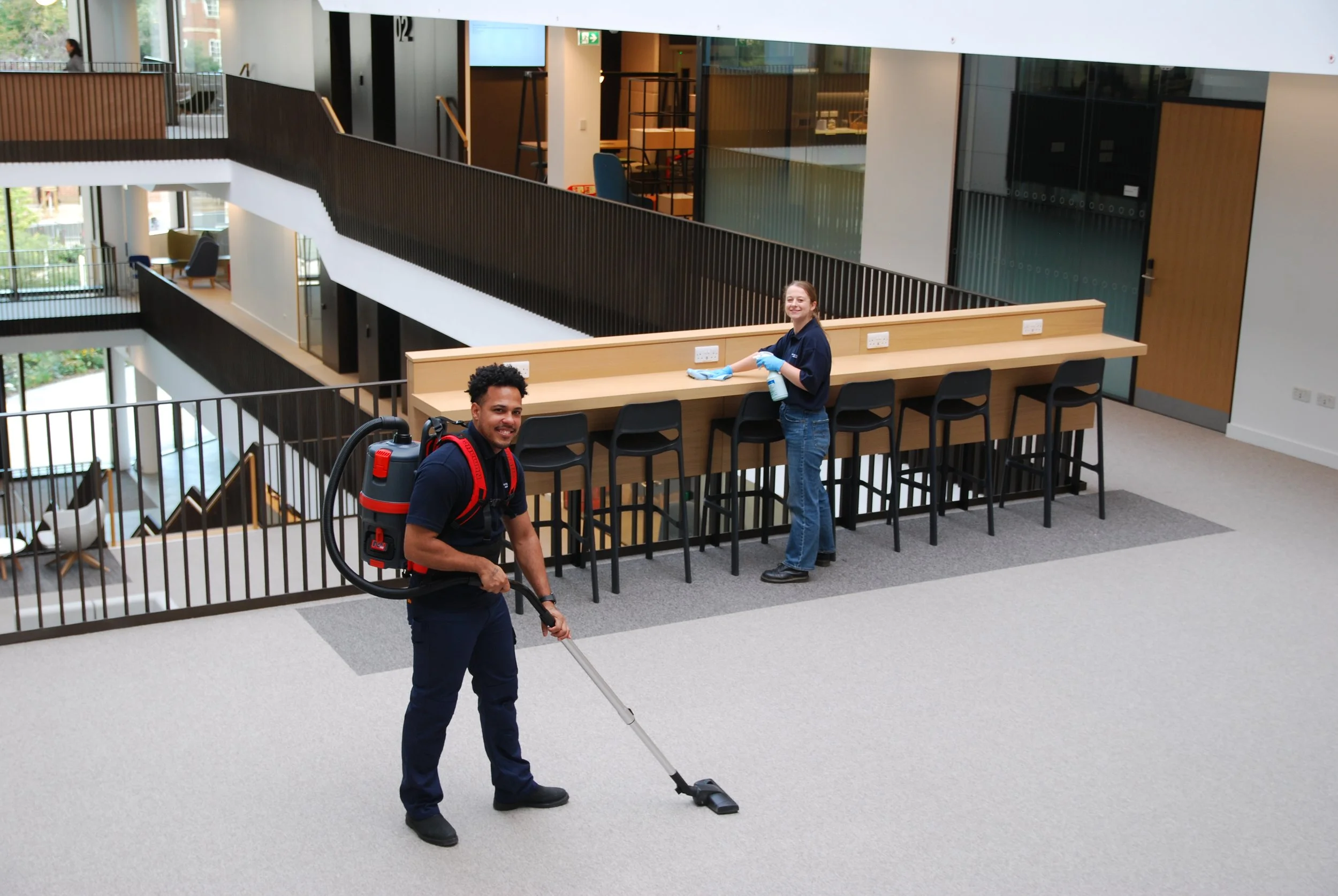 A man with a backpack vacuum cleaner and a woman in a navy shirt cleaning in a modern, open-concept office or public space with staircases and seating areas.